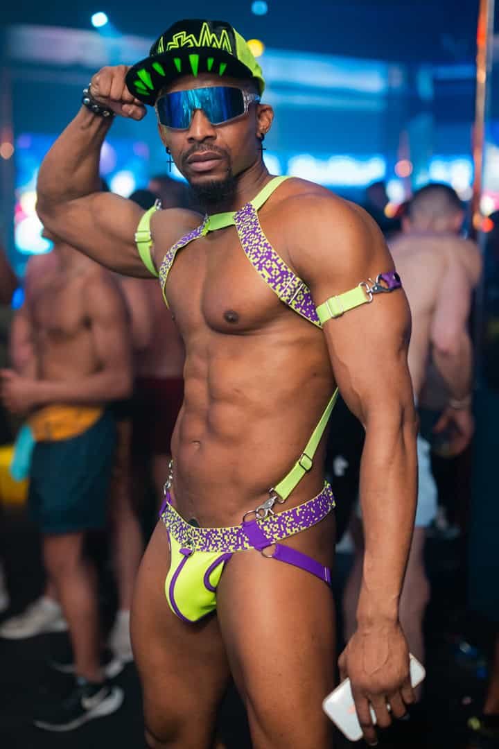 A man in colorful, revealing festival attire with neon green and purple details, wearing sunglasses.